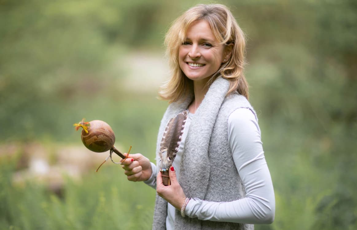 Smiling blonde woman in a grey vest holding a decorative gourd rattle and large feather.