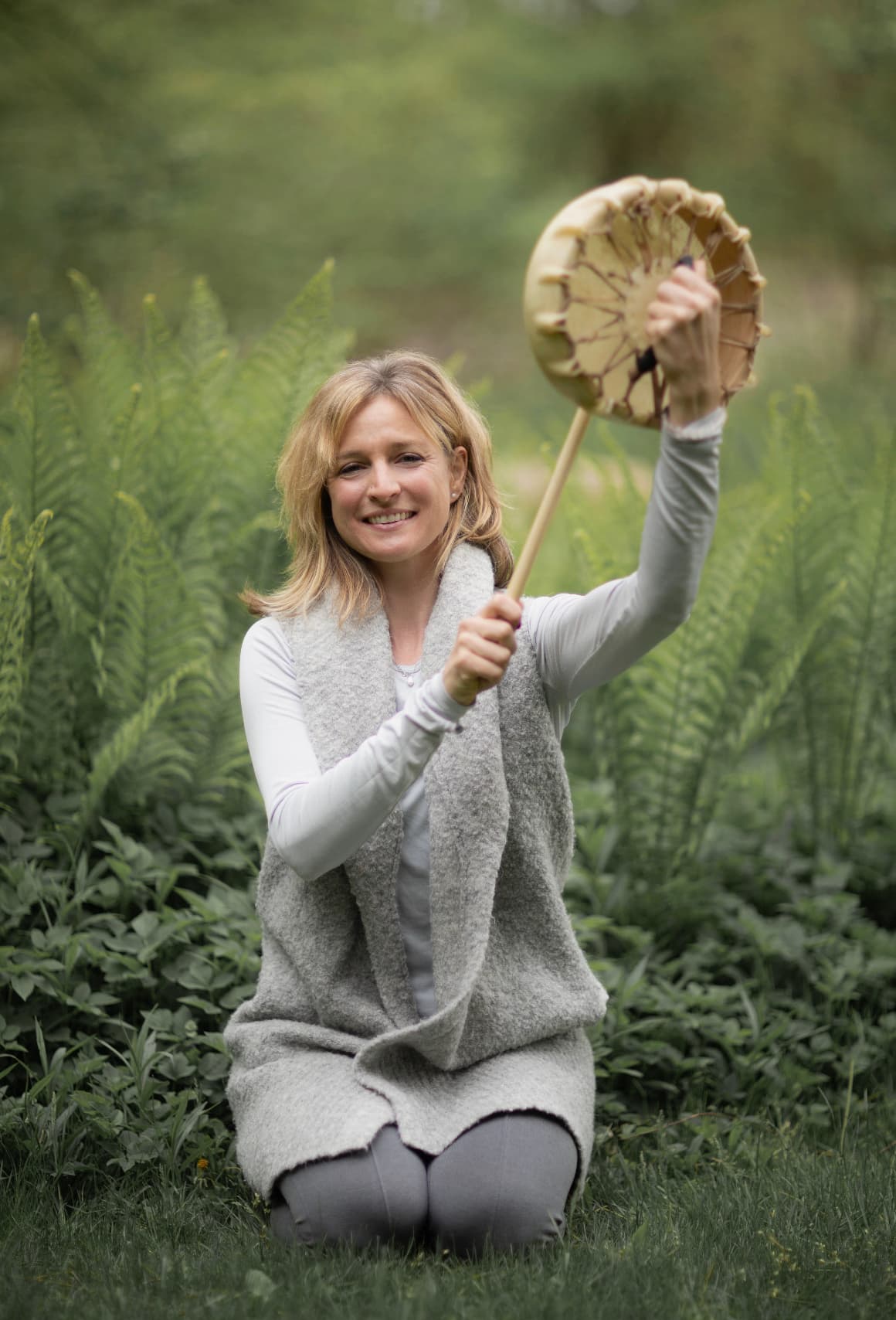 Smiling woman kneeling in lush ferns, holding a traditional frame drum and beater in nature.