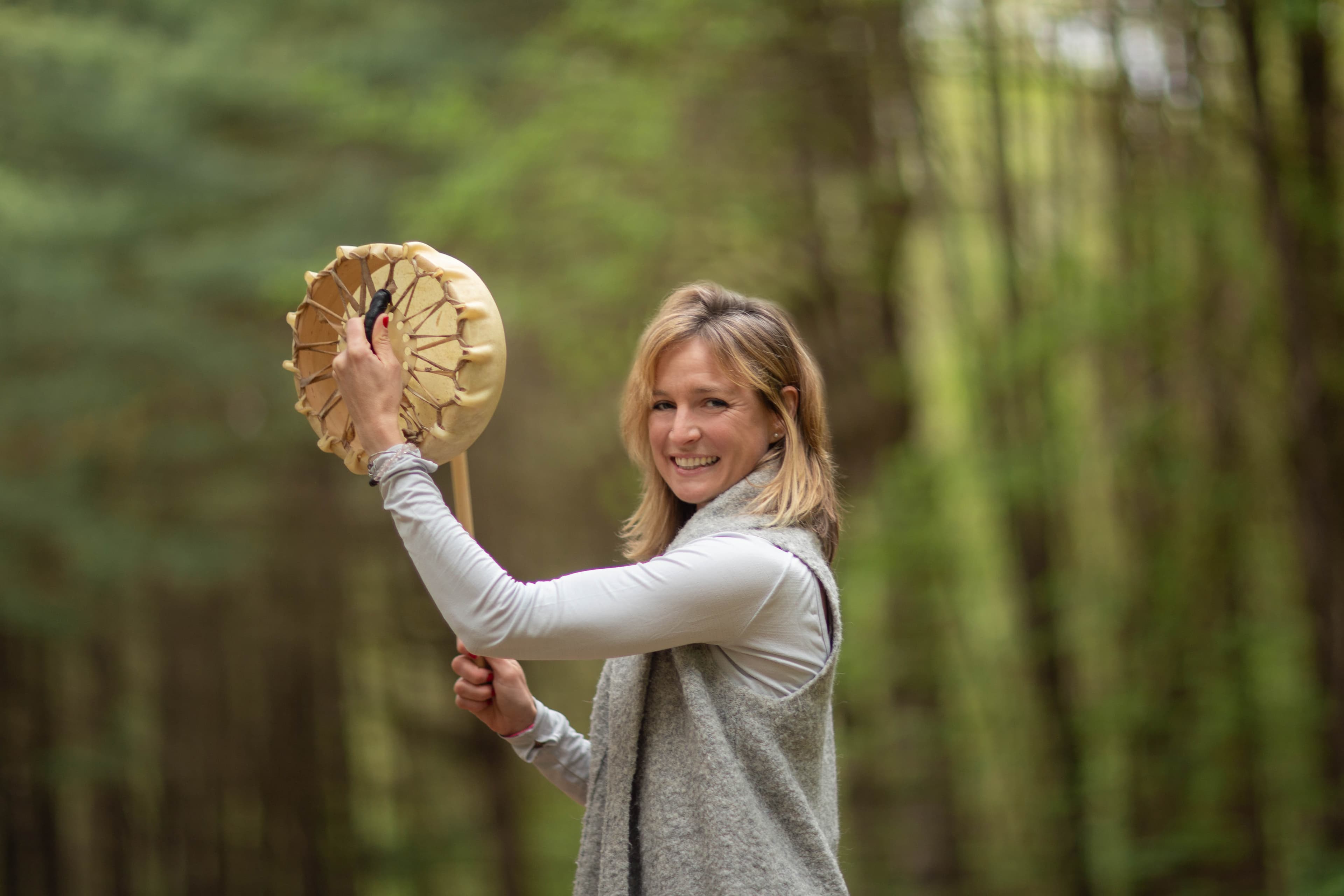 Smiling woman holding a traditional frame drum and mallet in a lush green forest.