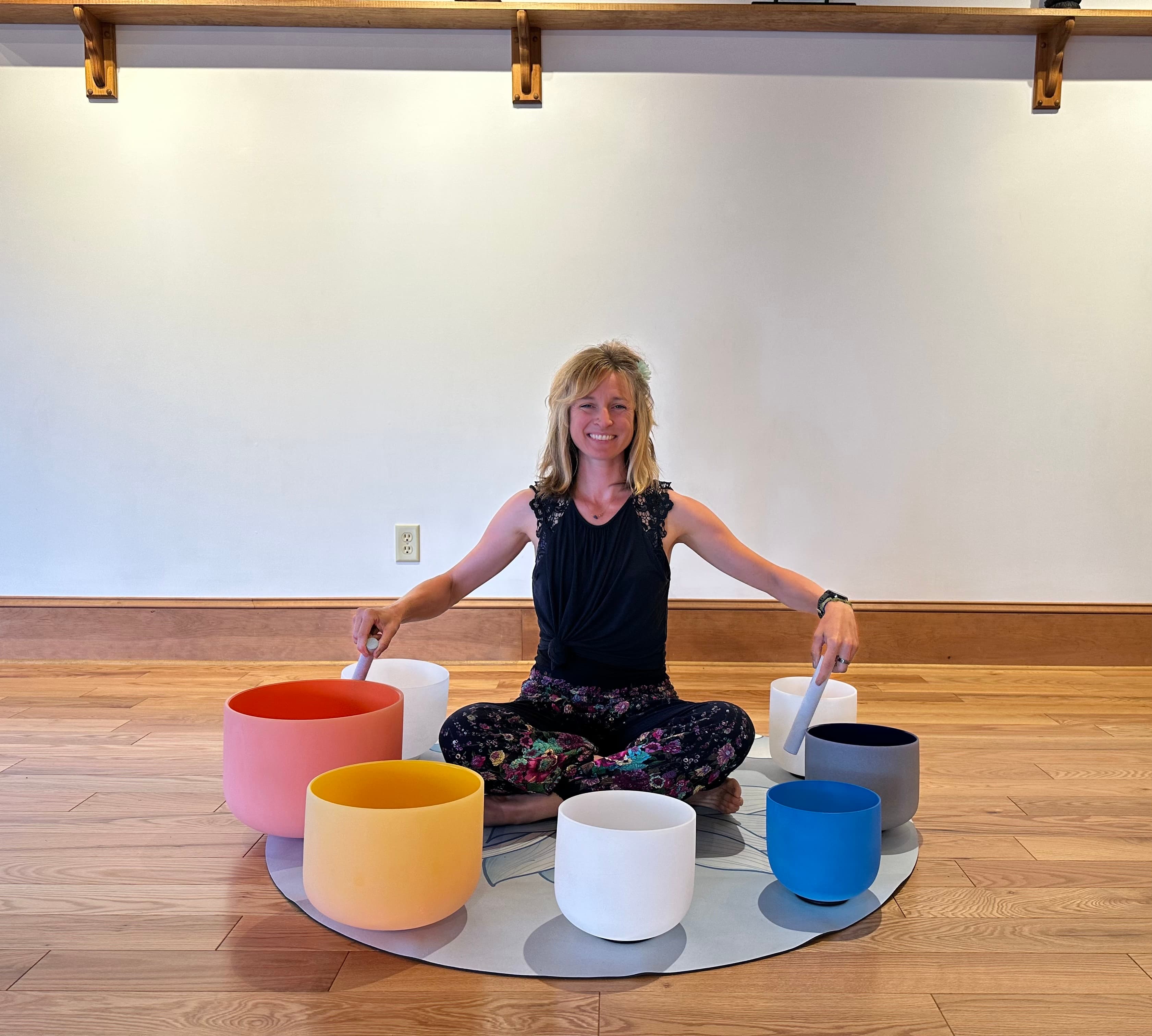 Smiling woman sits cross-legged on a mat, holding mallets over colorful crystal singing bowls.