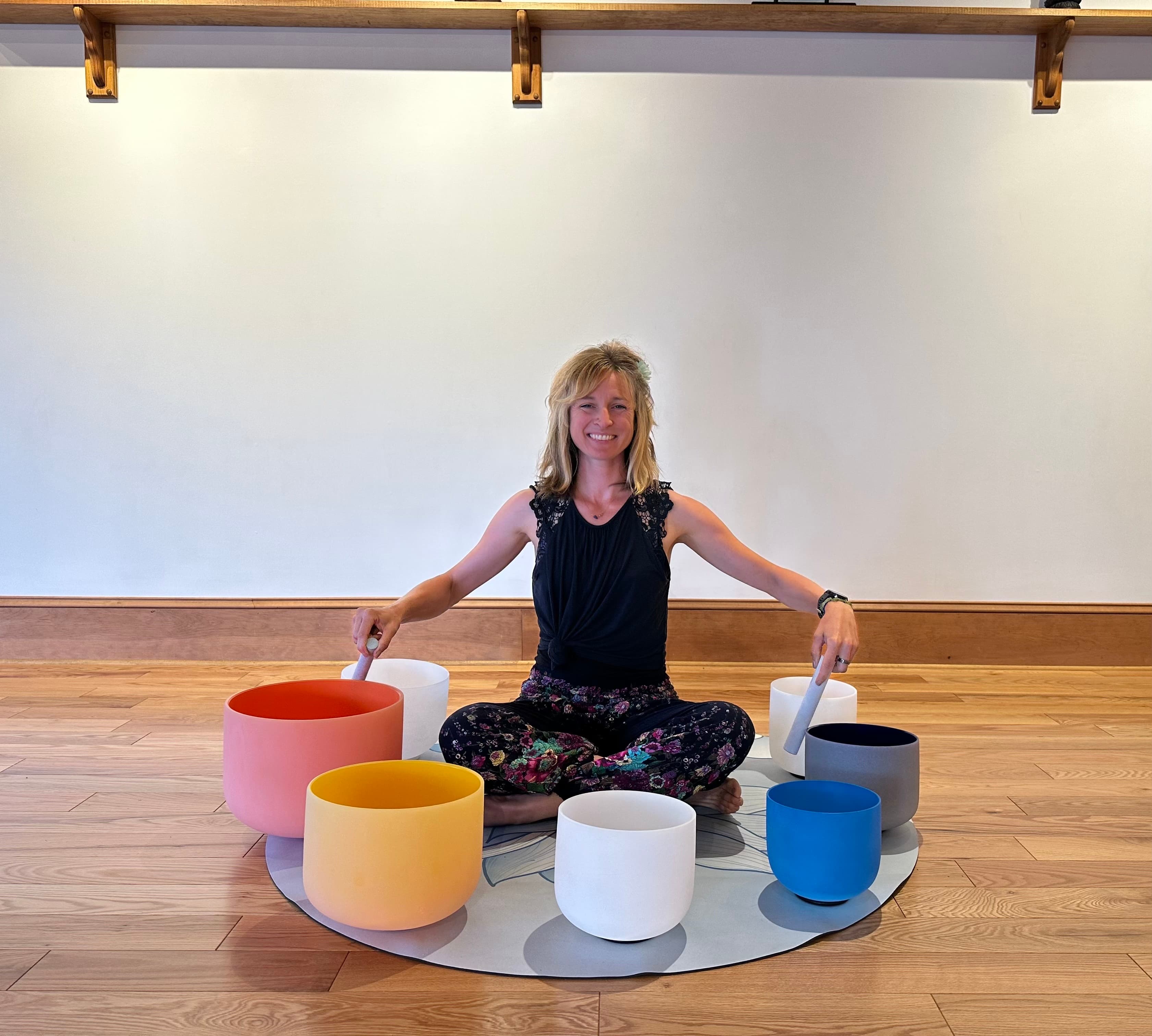 Smiling woman sits cross-legged on a mat surrounded by seven colorful crystal singing bowls.