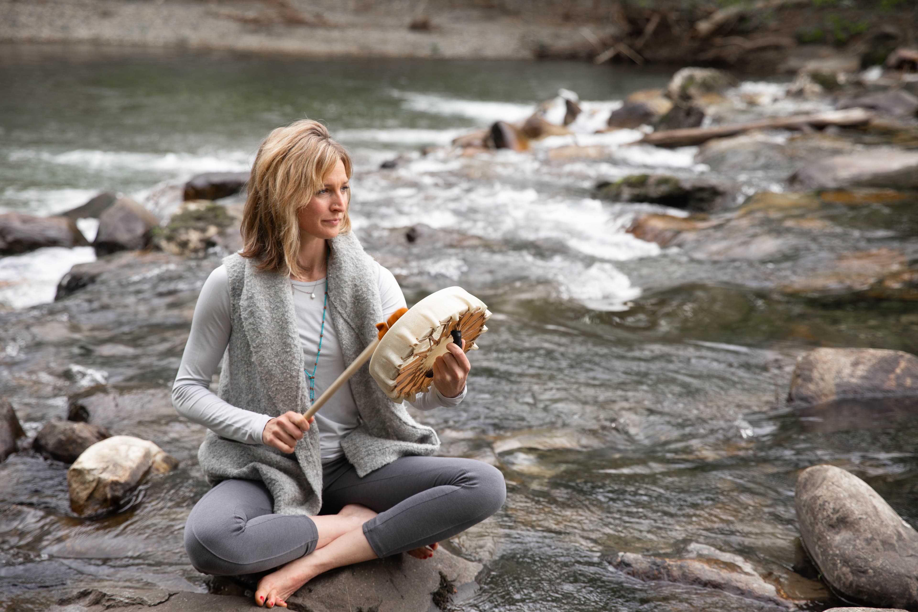Blonde woman sits cross-legged on a river rock, holding a frame drum and mallet.
