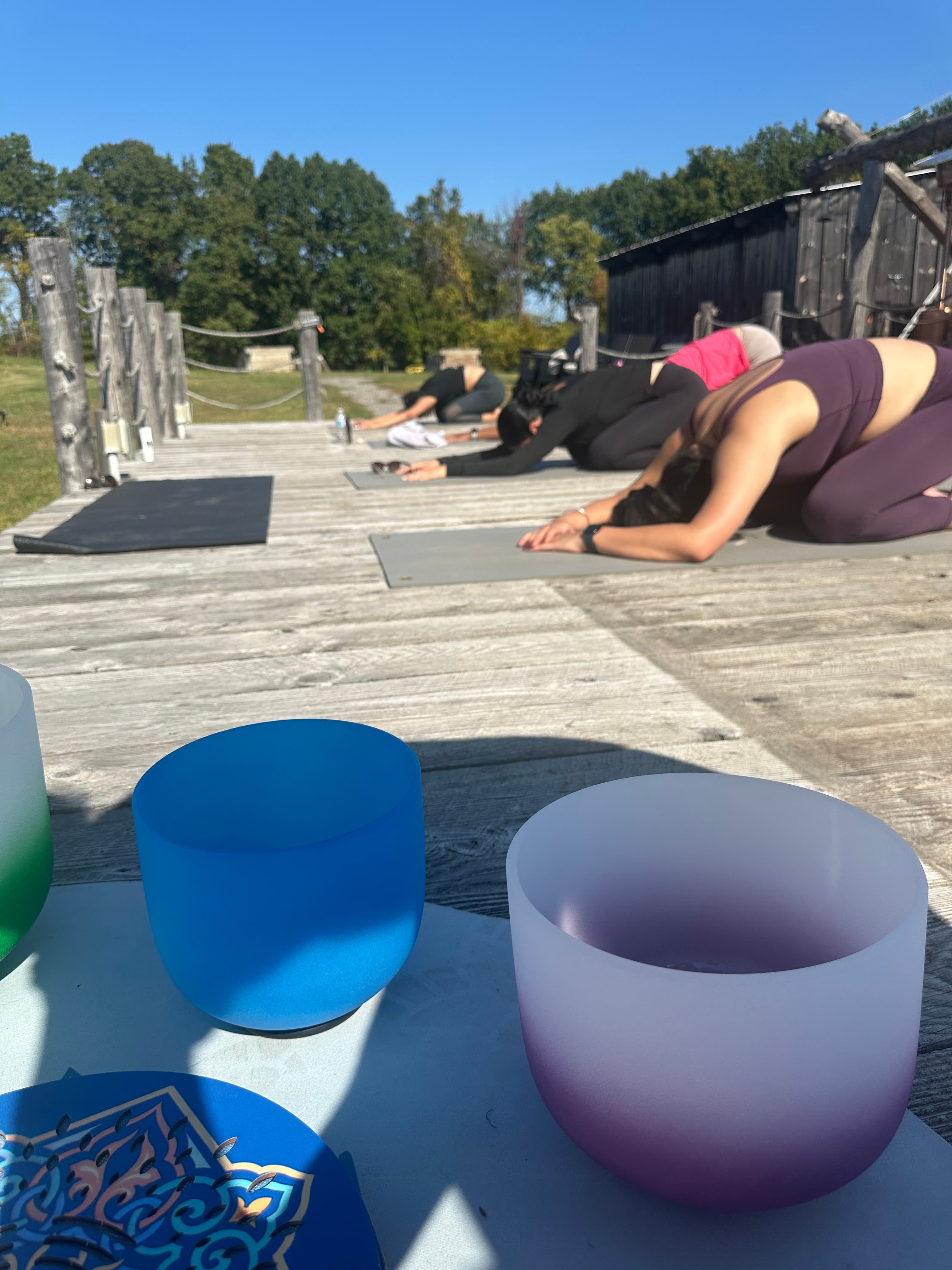 People practicing yoga in child's pose on a wooden deck behind colorful crystal singing bowls.